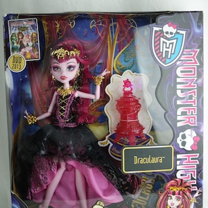 Op de afbeelding: Monster High Draculaura pop in een doos. De pop draagt roze en zwarte kleding, met een gouden tiara en sieraden. De doos bevat een dvd uit 2013 en de tekst "13 Wishes". De pop zit in een decoratieve doos met een rood accessoire.