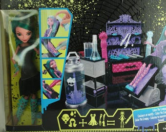 Peut inclure: Coffret de poupée Monster High. La boîte présente une poupée aux cheveux verts et une robe noire, ainsi qu'un ensemble de coiffeuse violet et rose. L'ensemble comprend des accessoires et le texte "Color Me Creepy".