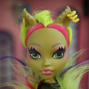 Peut inclure: Une poupée à la peau verte avec des yeux vert vif et des cheveux roses et violets. Elle porte un bandeau rose et a une paire de bois sur la tête.