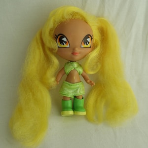May include: A small doll with long, yellow hair and large eyes. The doll is wearing a green crop top, a matching skirt, and green boots with yellow soles. The doll's skin is tan, and it has a small smile.
