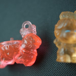 May include: Two small plastic figurines, one is red and the other is yellow. The red figurine is shaped like a small animal with a crown on its head. The yellow figurine is shaped like a person with a crown on its head.