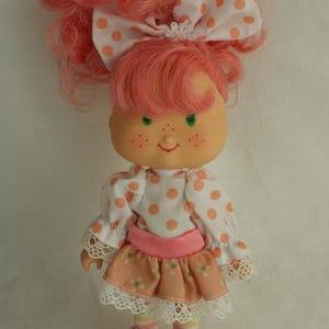 Könnte beinhalten: Eine Vintage-Puppe mit rosa Haaren, grünen Augen und Sommersprossen. Die Puppe trägt ein weißes Oberteil mit orangefarbenen Punkten, einen rosa Rock und rosa Schuhe. Eine große Schleife mit orangefarbenen Punkten ziert den Kopf der Puppe. Ein Sammlerstück.