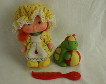 Puede incluir: Una muñeca amarilla y verde con un vestido amarillo y blanco de lunares y un sombrero amarillo y blanco. La muñeca sostiene una tortuga de juguete verde y roja.
