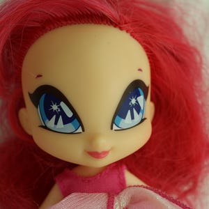 May include: Close-up of a doll's face with bright red hair and large blue eyes. The doll has a fair complexion, a small pink mouth, and is wearing a pink dress. The eyes have a star-like design.