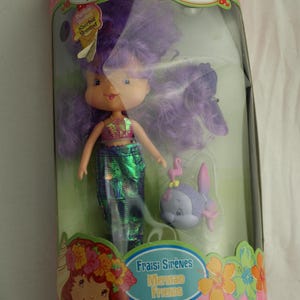 May include: A Strawberry Shortcake Mermaid Friends doll in a clear plastic package. The doll has purple hair, a pink and green top, and a green mermaid tail. A purple fish figure is also included. The packaging reads "Charlotte aux Fraises".