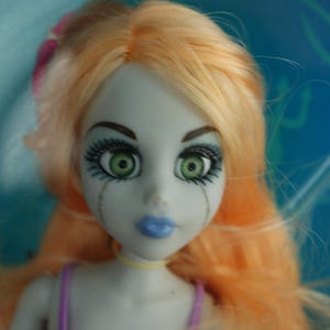 May include: A doll with pale gray skin, vibrant green eyes, and long peach-colored hair. The doll features dark eyelashes, blue lips, and thin vertical lines extending from the eyes down the cheeks. It wears a purple strap and a yellow choker.