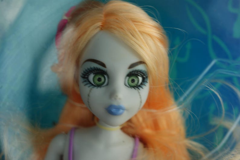 May include: A doll with pale gray skin, vibrant green eyes, and long peach-colored hair. The doll features dark eyelashes, blue lips, and thin vertical lines extending from the eyes down the cheeks. It wears a purple strap and a yellow choker.