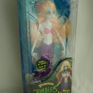 May include: A Zombie La Sirenita doll in original packaging. The doll has pale skin, long light orange hair, a pink seashell top, and a purple tail. The box features the text Zombie La Sirenita, Princesas ZOMBIES, and La Sirenita A Pequena Sereia.