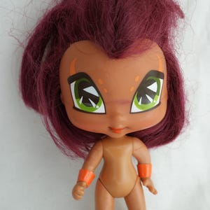 May include: A small doll with a tan complexion, large green eyes, and dark red hair. The doll has orange armbands and a small orange mouth. The doll is standing on a white surface.
