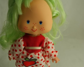 Può includere: Una bambola con capelli verde chiaro e un accessorio rosso, che indossa un vestito a pois rossi e bianchi con un disegno di una mela sul davanti. La bambola ha leggings a righe, scarpe rosse e un viso dipinto con occhi azzurri e lentiggini.