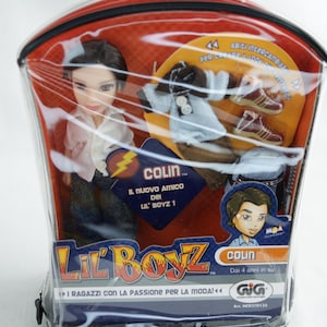 Könnte beinhalten: Eine Lil' Boyz-Puppe namens Colin in einer transparenten Plastikverpackung. Die Puppe trägt ein rosa Hemd und Jeans. Die Verpackung enthält zusätzliche Kleidung und Schuhe. Auf der Verpackung steht "Colin, Il Nuovo Amico Dei Lil' Boyz!" und "I Ragazzi Con La Passione Per La Moda!"
