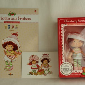 Op de afbeelding: Een Strawberry Shortcake pop in een rode doos, een boekje getiteld Charlotte aux Fraises met een karakterillustratie, en een kleine rechthoekige kaart met twee Strawberry Shortcake figuren. De pop draagt een witte hoed, een poncho met patroon en groen gestreepte leggings.