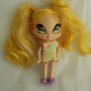 May include: A small doll with yellow hair styled in pigtails. The doll has large eyes and is wearing a yellow dress with pink trim and purple shoes. The dress has a small floral design on the front.