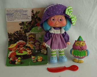 May include: A purple and blue Strawberry Shortcake doll with a green and white bow, wearing a purple dress with white lace trim and white stockings. The doll is holding a pink owl with green wings and a red accordion.