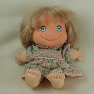 May include: A smiling doll with blonde hair wearing a floral dress. The doll has blue eyes and a pink and white collar.