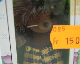 May include: A brown and gray hedgehog plush toy wearing a yellow and black plaid shirt. The toy is in a clear plastic box with a green background. The box has a price tag that says "085 Fr. 150".