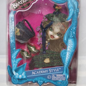 Könnte beinhalten: Bratzillaz Academy Style Puppe in einer transparenten Kunststoffverpackung. Die Puppe hat grüne Augen, rote Lippen und lockiges braunes Haar. Sie trägt ein grünes Kleid und einen Hut. Zubehör: schwarze Handtasche und grüne Schuhe. Die Verpackung hat einen blauen dekorativen Rahmen.