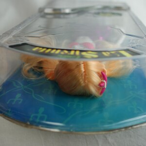 May include: A doll with long blonde hair and a small pink hair accessory, encased in a clear plastic blister pack. The packaging features a blue cardboard backing with a repeating trident pattern and the text La Sirenita printed in yellow.