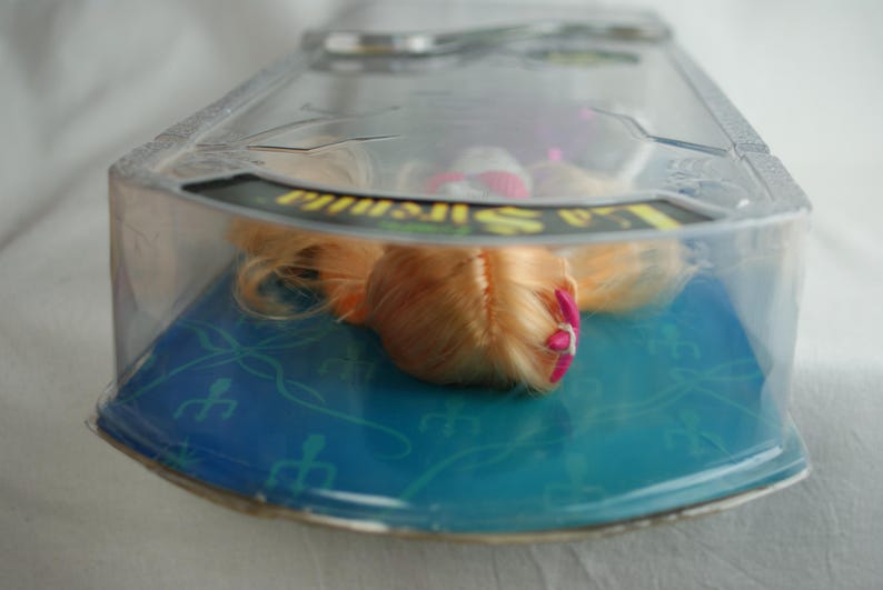 May include: A doll with long blonde hair and a small pink hair accessory, encased in a clear plastic blister pack. The packaging features a blue cardboard backing with a repeating trident pattern and the text La Sirenita printed in yellow.