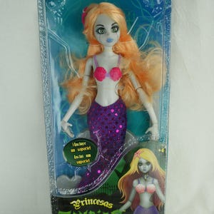 May include: A La Sirenita Zombie doll in original packaging with long orange hair, pale skin, and a purple sequined mermaid tail. The doll wears a pink seashell top. The box features the text La Sirenita, Princesas Zombies, and Famosa.