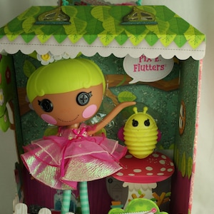 May include: A Lalaloopsy doll in a house-shaped box. The doll has button eyes, pink cheeks, and a pink dress. A yellow bug sits on a red mushroom. The box has a green roof and a white picket fence.