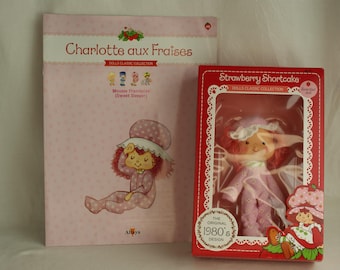 Può includere: Un libro rosa intitolato "Charlotte aux Fraises" con un'illustrazione di un personaggio dei cartoni animati in pigiama. Accanto, una bambola Strawberry Shortcake in scatola degli anni '80, in una scatola rossa con finestra trasparente.