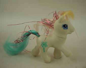 May include: A white My Little Pony toy with a blue and pink mane and tail. The pony has a blue eye with a white star and a turquoise symbol on its flank.