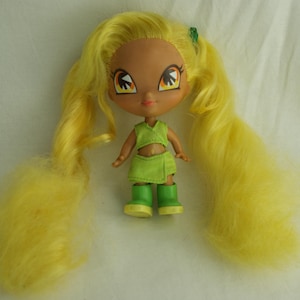 Puede incluir: Muñeca con pelo largo y amarillo en coletas. La muñeca tiene tez bronceada, ojos grandes y viste un atuendo verde con botas a juego. Un pequeño accesorio verde está en el pelo.