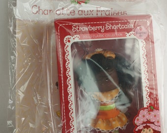 Puede incluir: Una caja roja con un borde blanco y una imagen de una muñeca Strawberry Shortcake. La muñeca lleva un vestido amarillo y naranja con lunares blancos. La caja dice "Strawberry Shortcake Dolls Classic Collection" y "The Original 1980's Design".