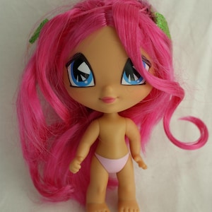 Może przedstawiać: Stojąca lalka z jasnoróżowymi włosami i dużymi niebieskimi oczami. Lalka ma opaloną cerę i nosi jasnoróżowe majtki od bikini. Zielone akcenty są widoczne we włosach. Włosy lalki są długie i falowane.