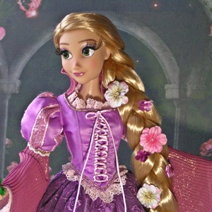 May include: A doll of a princess with long blonde hair adorned with flowers. She wears a purple dress with pink sleeves and a white lace-up bodice. A small green frog is held in her hand. The background features a rose garden.