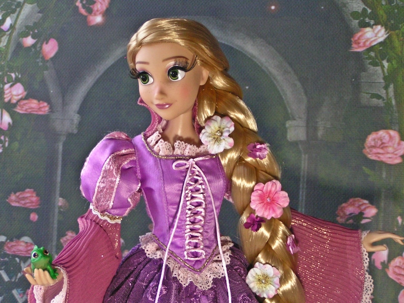 May include: A doll of a princess with long blonde hair adorned with flowers. She wears a purple dress with pink sleeves and a white lace-up bodice. A small green frog is held in her hand. The background features a rose garden.