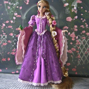 May include: A doll of a princess in a long purple gown with a corset-style bodice and long blonde hair adorned with flowers. The dress has a lace trim and is embellished with embroidered roses. The doll is holding a small green frog.