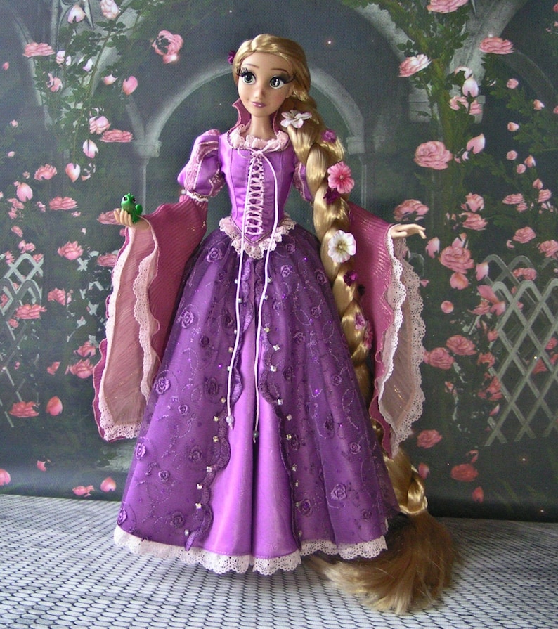 May include: A doll of a princess in a long purple gown with a corset-style bodice and long blonde hair adorned with flowers. The dress has a lace trim and is embellished with embroidered roses. The doll is holding a small green frog.