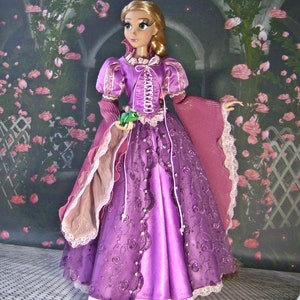 May include: A doll dressed in a purple gown with a corset-style bodice and lace trim. The doll has long blonde hair and is holding a small green frog. The background features a floral archway.