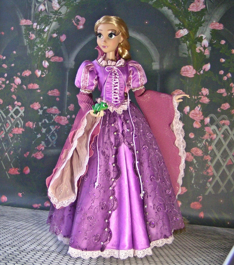 May include: A doll dressed in a purple gown with a corset-style bodice and lace trim. The doll has long blonde hair and is holding a small green frog. The background features a floral archway.