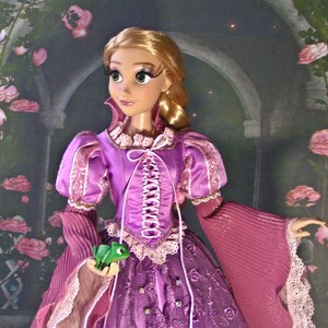 May include: A Rapunzel doll with long blonde hair, wearing a purple dress with pink accents and a lace-up bodice. The doll holds a small green chameleon. The dress features puffed sleeves and a long skirt with embroidered details.
