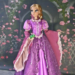 May include: A doll of a princess in a long, purple gown with a corset-style bodice and intricate lace details. The doll has long blonde hair and is holding a small green frog. The background features a floral design.