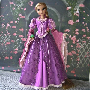 May include: A doll dressed in a purple gown with a corset-style bodice and long sleeves. The dress features lace and floral embellishments. The doll has long blonde hair and is holding a small green frog.