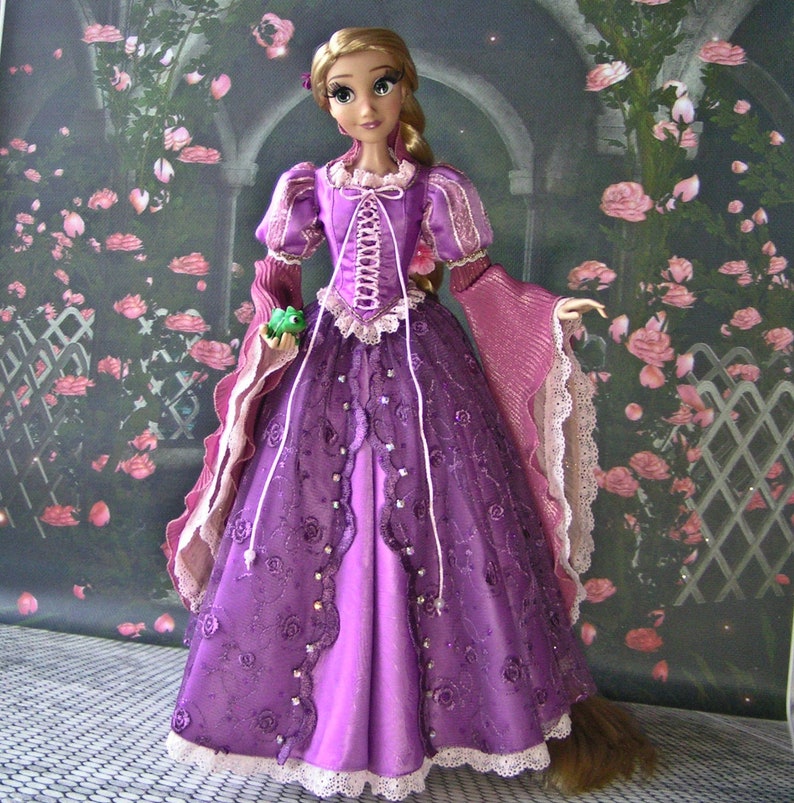 May include: A doll dressed in a purple gown with a corset-style bodice and long sleeves. The dress features lace and floral embellishments. The doll has long blonde hair and is holding a small green frog.