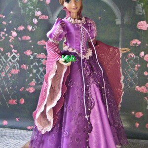 May include: A doll wearing a long, purple gown with intricate lace and floral embroidery. The doll has long blonde hair and is holding a small green frog. The gown features a high collar and long, flowing sleeves.