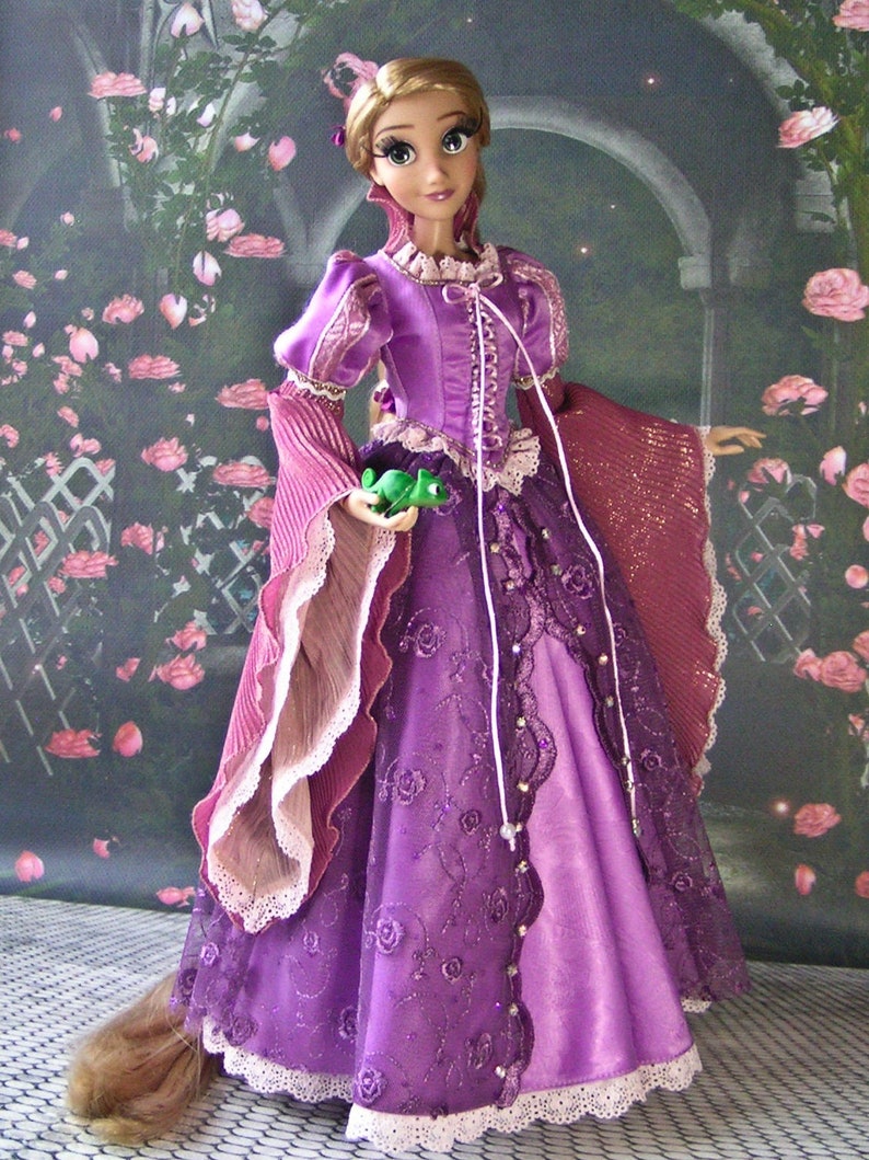 May include: A doll wearing a long, purple gown with intricate lace and floral embroidery. The doll has long blonde hair and is holding a small green frog. The gown features a high collar and long, flowing sleeves.
