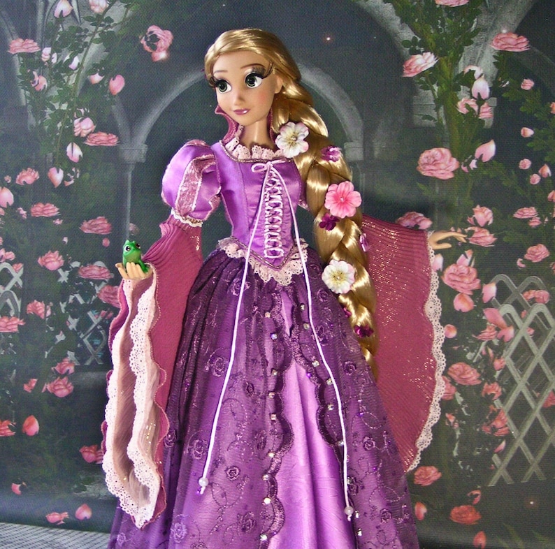 May include: A doll dressed in a purple gown with a corset-style bodice and long, flowing sleeves. The doll has long blonde hair adorned with flowers and is holding a small green frog. The background features a floral design.