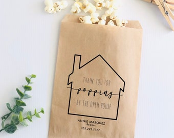 May include: A brown paper bag with a house-shaped design and the text "Thank you for popping by the open house."  Angie Marquez Realtor 303 222 7777 is printed on the bag. The bag is filled with popcorn.