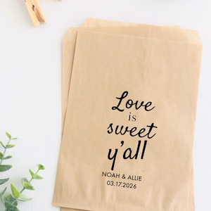 May include: Two brown paper favor bags with the text "Love is sweet y'all" in black script. The names "Noah & Allie" and the date "03.17.2026" are printed below. Two wooden clothespins and greenery are in the background.