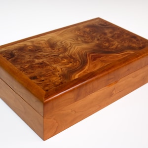 May include: A wooden box with a natural wood finish. The top of the box is made of a different type of wood with a unique grain pattern. The box is closed.