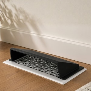 May include: A rectangular floor vent cover featuring a matte black finish with a slanted hood design. The interior grate displays a repeating geometric pattern of intersecting lines and squares, set against a white base plate on a wooden floor.