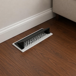 May include: A rectangular floor register vent featuring a white base with a decorative geometric lattice pattern and a black metal deflector hood attached to the top. The vent is positioned on a dark brown wood floor against a white baseboard.