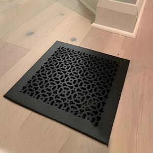 以下が含まれることがあります： マットブラックの金属製スクエア型フロアレジスターカバー。円形と十字形が組み合わさった幾何学的な格子模様が繰り返されており、明るい色の木製床面にフラットに収まるように設計されています。
