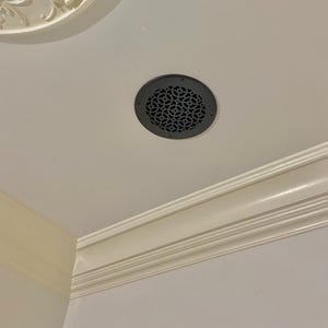 May include: A black round ceiling vent with a decorative, intricate pattern. The vent is installed in a white ceiling with white crown molding.